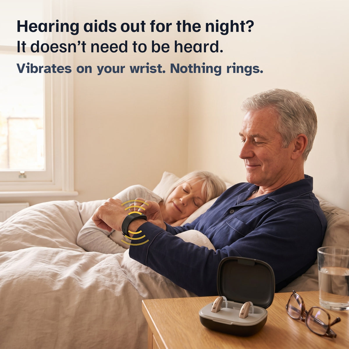 Open hearing-aid case on a bedside table while a man in his 60s checks the band on his wrist and his wife sleeps. Hearing aids out for the night? It doesn’t need to be heard.