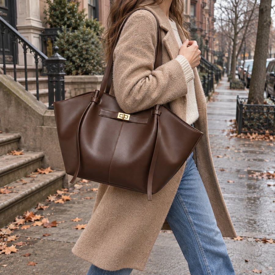 The Vela Tote in Chocolate, carried on the shoulder over a long camel coat, on an autumn street